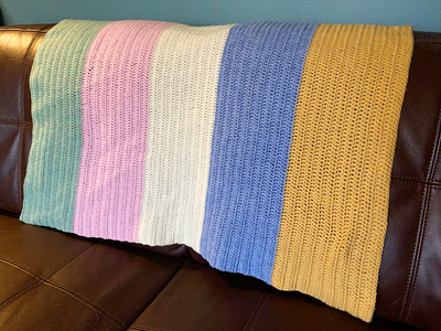 A crochet baby blanket in stripes of green, pink, cream, blue and yellow is draped over the back of a brown sofa