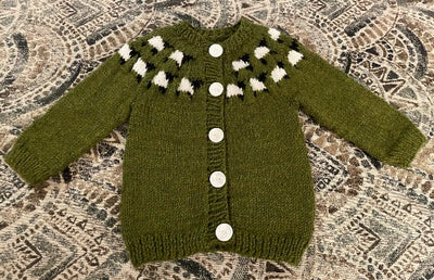 A green knitted baby cardigan with white buttons has a design of black and white sheep around the yoke