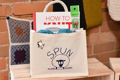 A cream canvas bag with rope handles contains red yarn, a book and leaflet and a pair of knitting needles