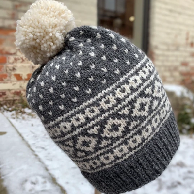 A charcoal grey knitted hat with white stitch patterns and a white pom pom