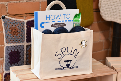 A cream canvas bag with rope handles contains skeins of cream yarn, a book and a crochet hook
