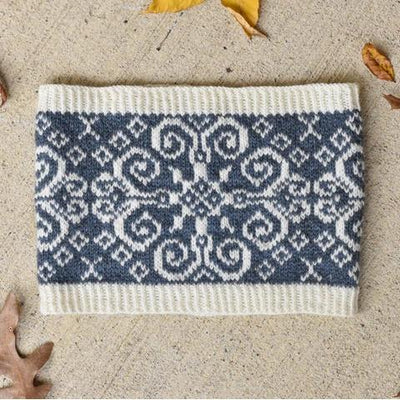 A cream and blue patterned knitted cowl on a cream background with autumn leaves