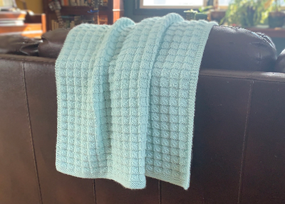 A mint green knitted baby blanket draped over the back of a dark brown leather couch
