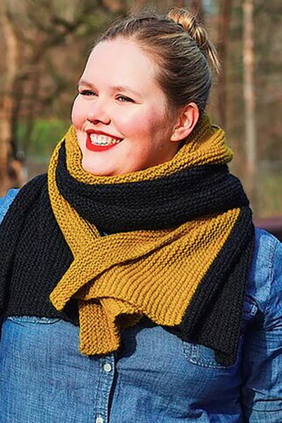 A blond woman wears a gold and dark grey knitted wrap over a blue denim shirt