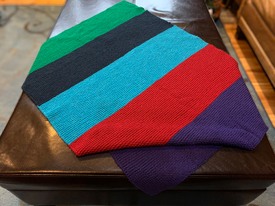 A knitted baby blanket with stripes of green, navy, turquoise, red and purple is spread on a dark brown table