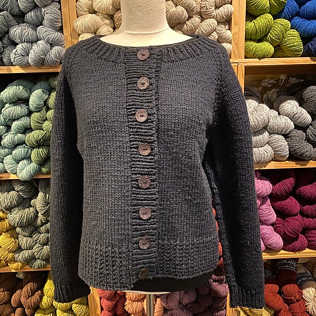 Photo of the Ramona Cardigan against a backdrop of colorful yarn on shelves. Cardigan is a simple design in dark blue with a crew neck, wide ribbing, and large buttons.