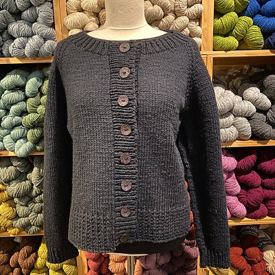 Photo of the Ramona Cardigan against a backdrop of colorful yarn on shelves. Cardigan is a simple design in dark blue with a crew neck, wide ribbing, and large buttons.